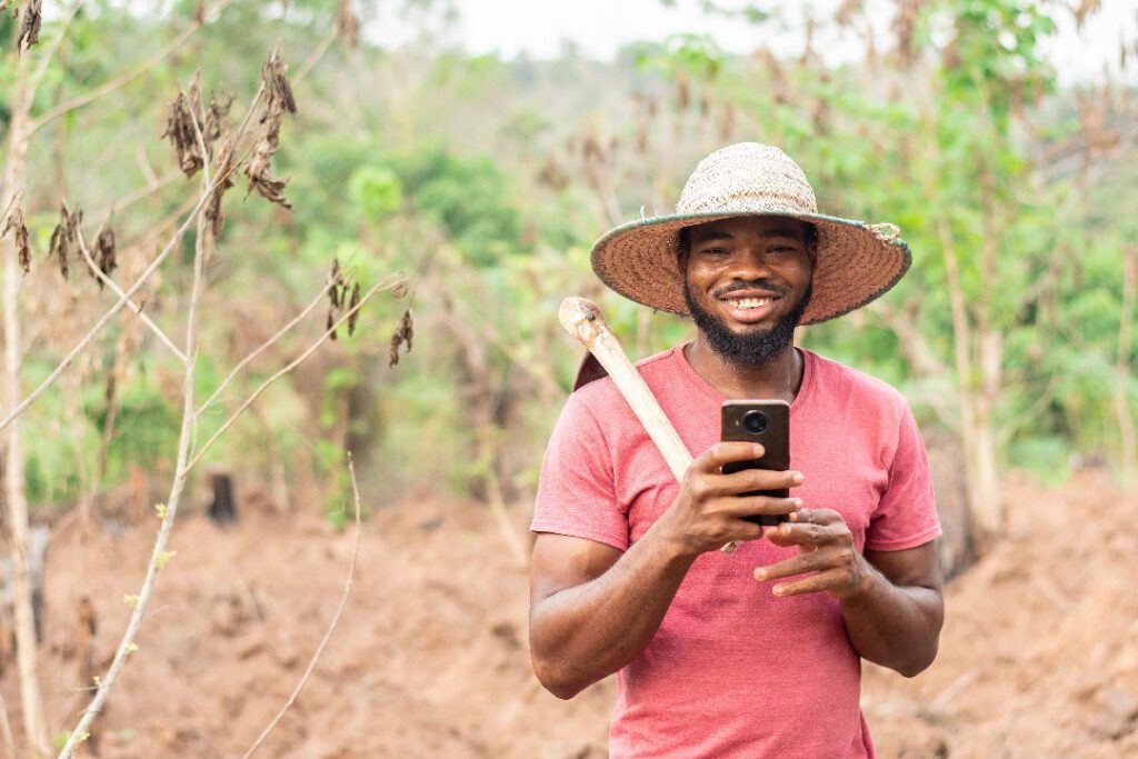 Smiling farmer in a straw hat holding a smartphone and hoe in a rural field, representing digital empowerment in agriculture